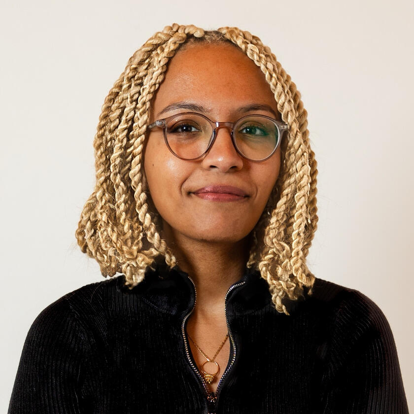 Photo of Edom, a Black woman with light skin and blonde twists, wearing glasses and a black turtleneck.